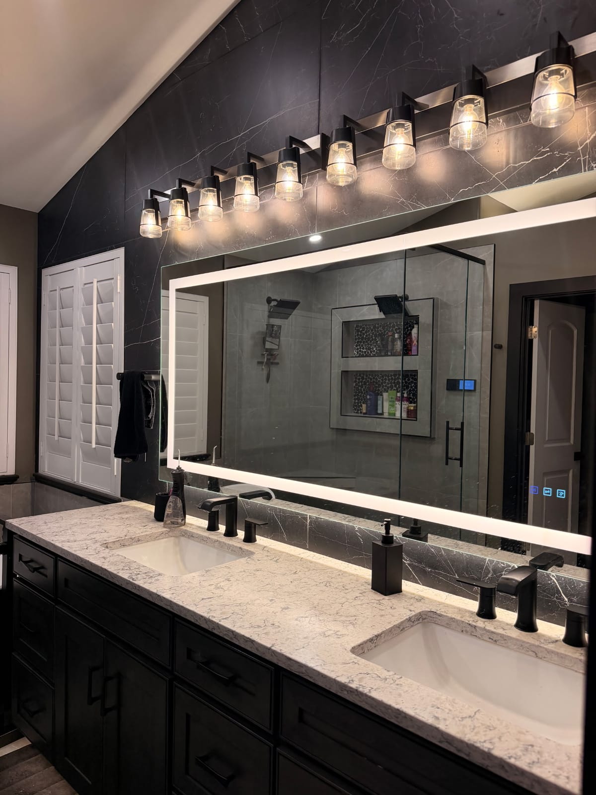 Double vanity in near-black shaker cabinetry with a light quartz countertop, two undermount basins and matte black faucets, below a wide backlit LED mirror and a row of black vanity lights with clear glass shades, against a black marble-look tiled wall.
